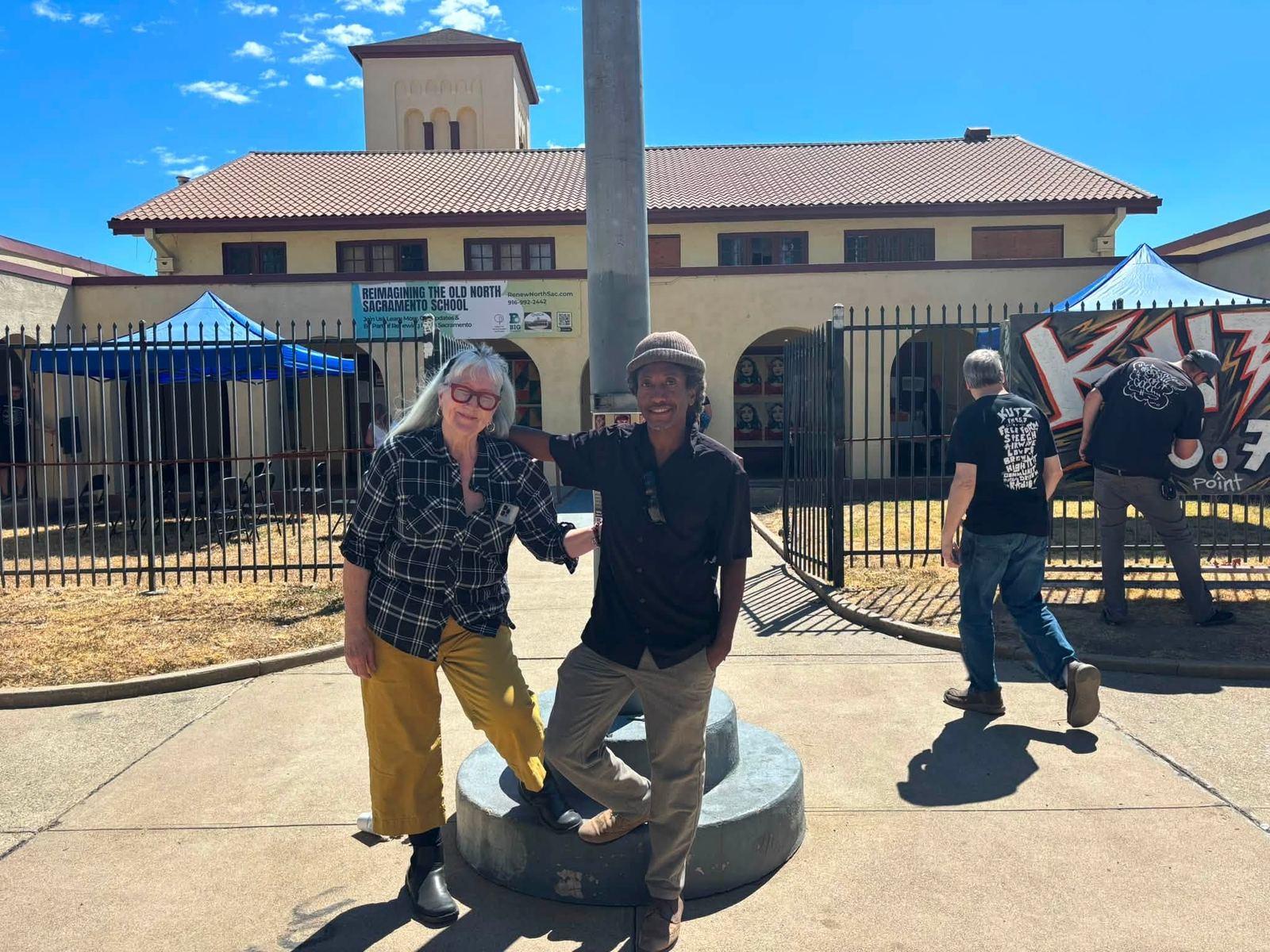 Kim Scott and Ronnie Shaw of the Old North Sacramento Community Association at the school's open front gate on Dixieanne Avenue, with tents on the lawn and the bell tower behind